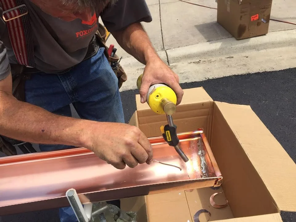 A man welding a copper pipe with a handheld welding tool, standing next to a cardboard box on the ground in a driveway.