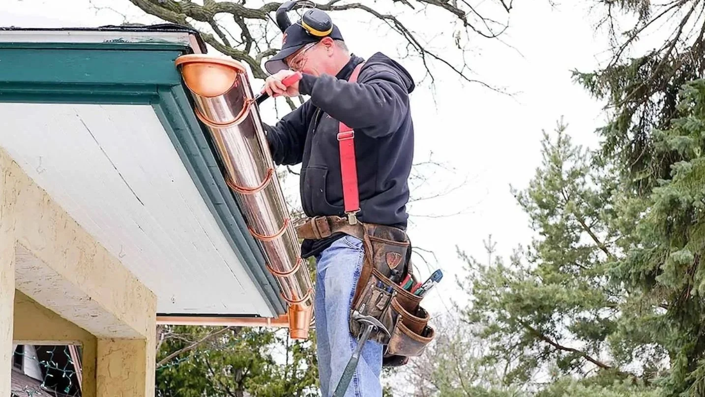 A man installing or repairing a copper gutter system. He is wearing a black hoodie, blue jeans, red suspenders, and a tool belt. He is also wearing ear protection.