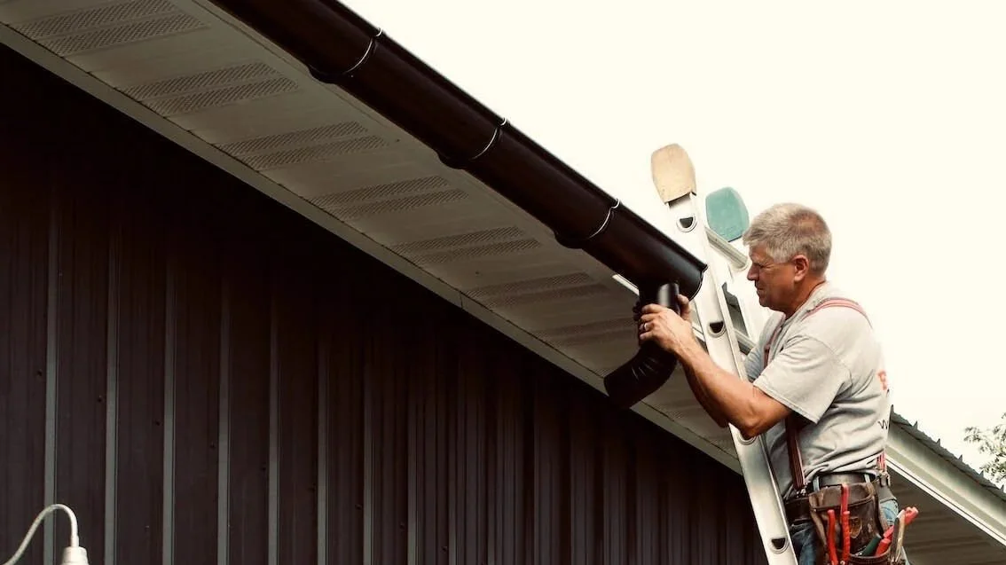 A man standing on a ladder working on a round downspout on a shed