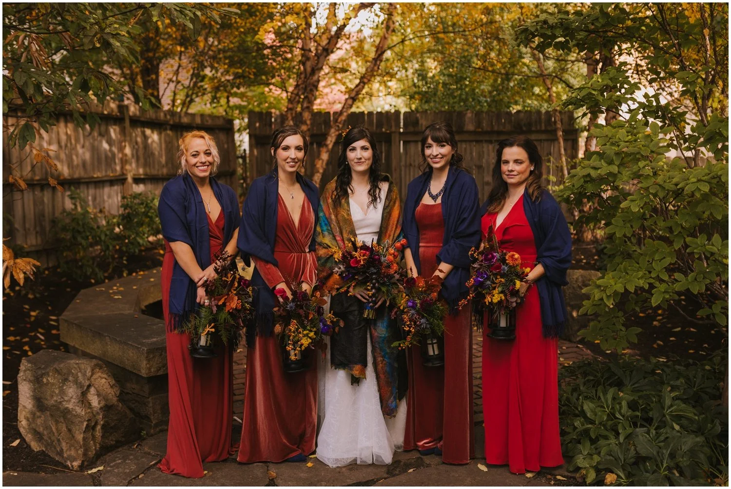 A bride and bridesmaids in orange bridesmaids dresses show off their colorful shawls in a Pittsburgh garden.