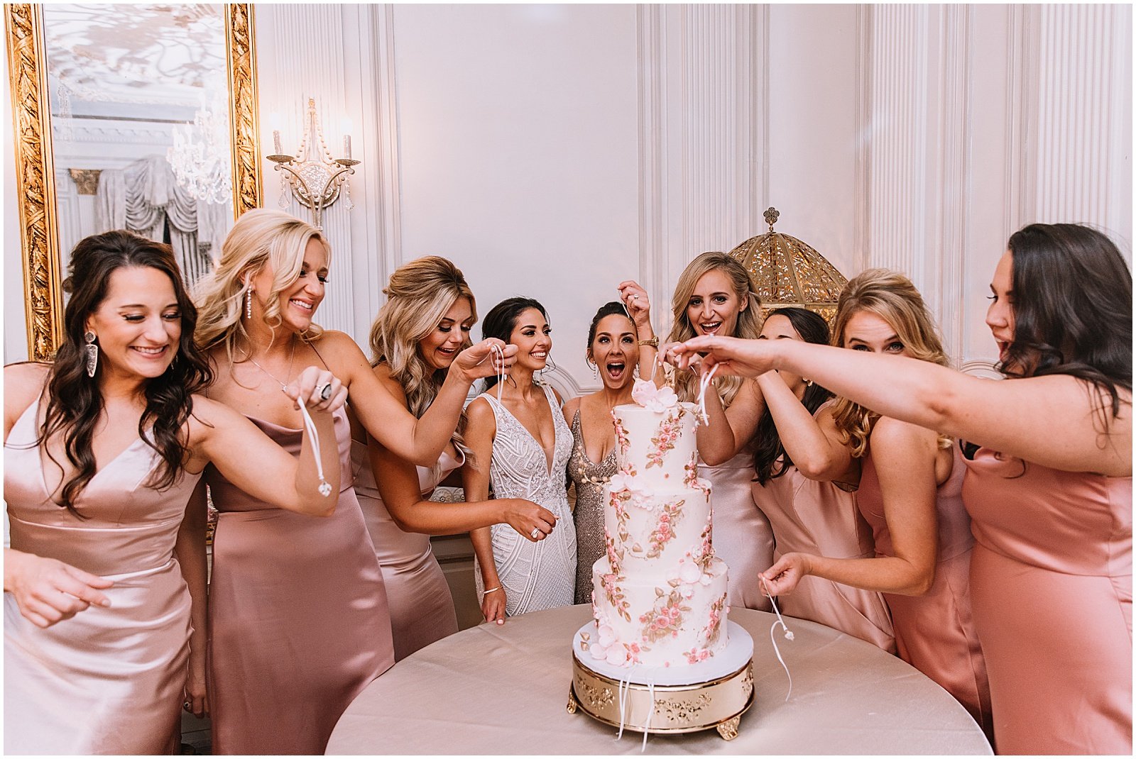 Bridesmaids gather around a thee-tiered wedding cake for a cake pull.