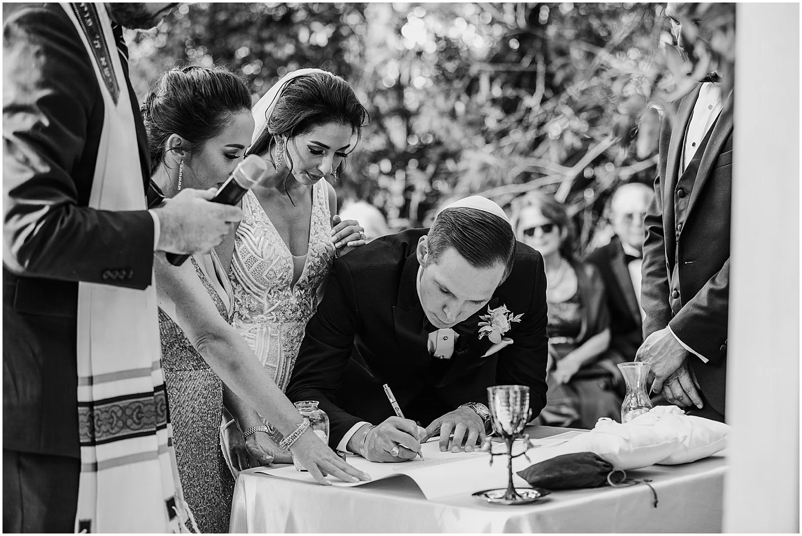A bride and groom lean over a table signing a ketubah during a Jewish wedding ceremony.