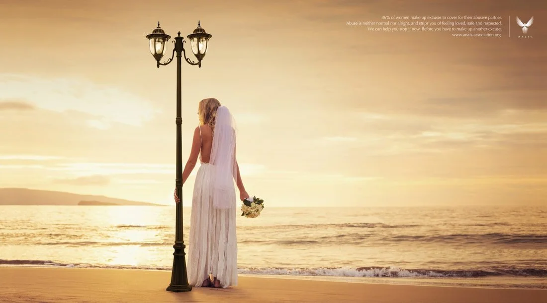 A woman in a white wedding dress standing on a beach at sunset, holding a bouquet, and leaning against a street lamp post.