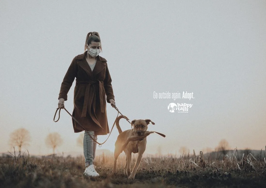 A woman wearing a face mask and a brown coat walks a dog on a leash in an open field during sunset or sunrise. Text on image reads: 'Go outside again. Adopt.' with Happy Tails logo.