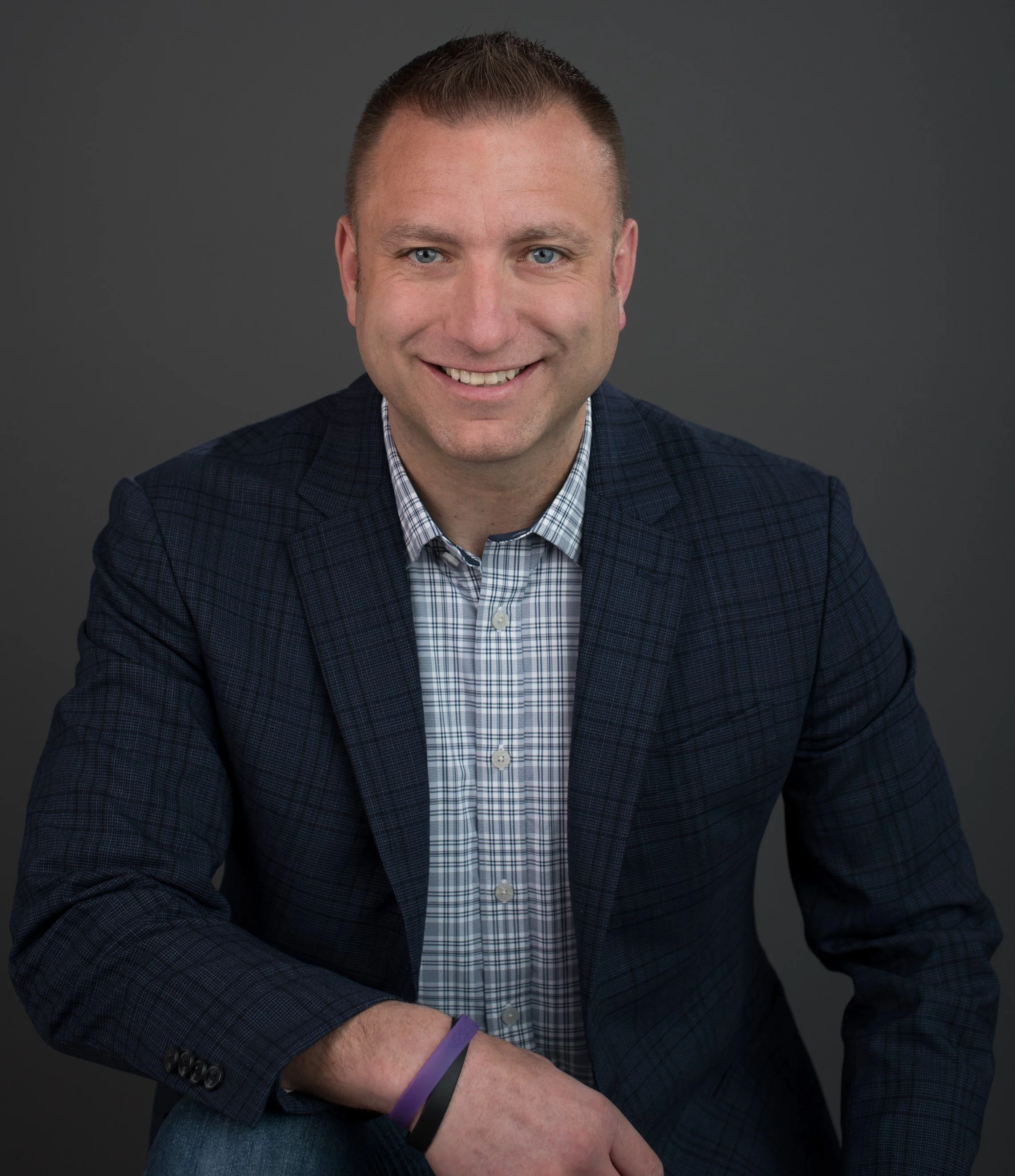 A smiling man with short brown hair and blue eyes, wearing a dark blue blazer, a checkered shirt, and a purple bracelet, sitting against a plain gray background.