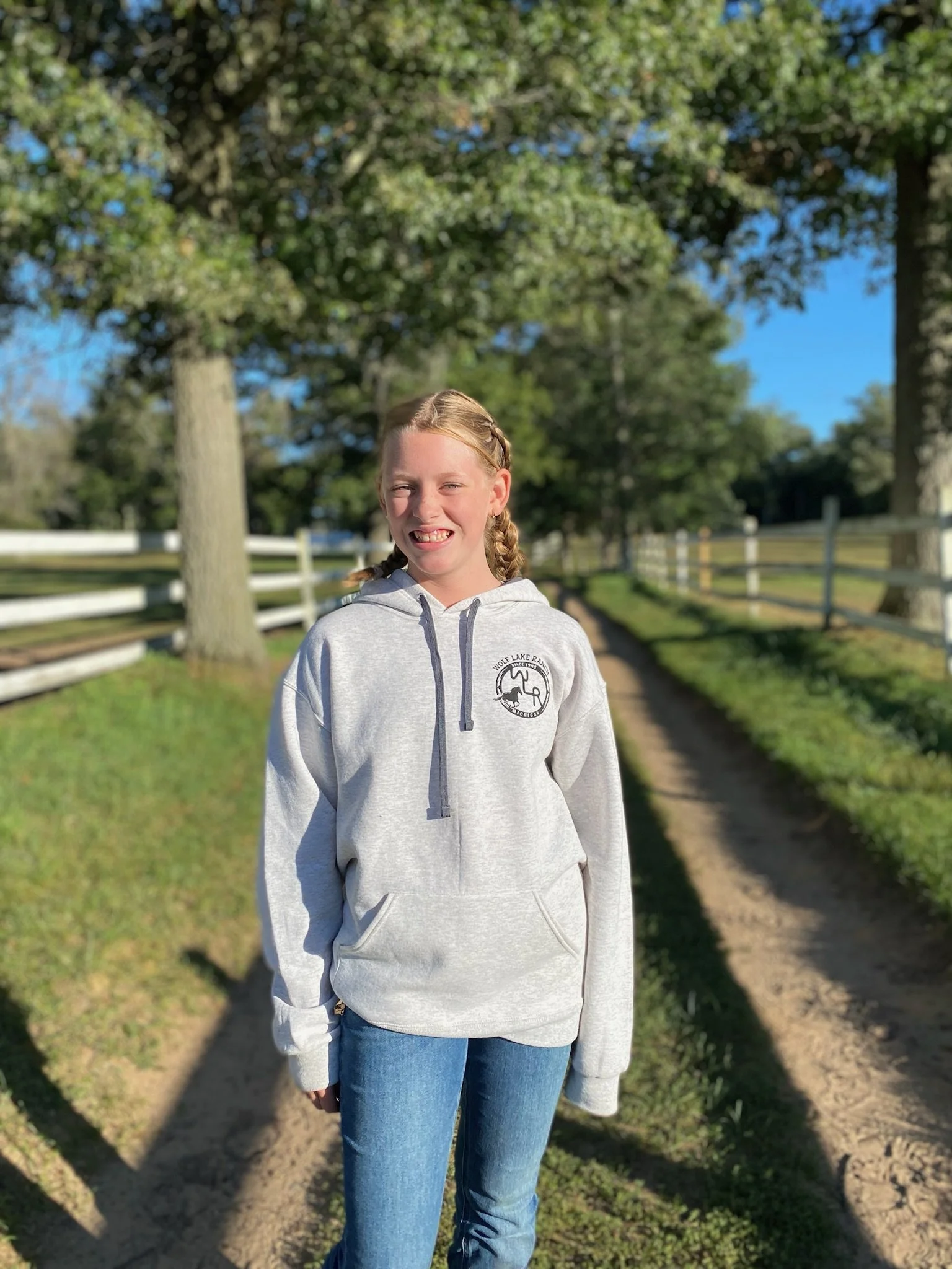 A young girl with blonde hair tied in braids, smiling and wearing a light gray hoodie with a logo on the chest, standing outdoors on a dirt path with green trees and blue sky in the background.