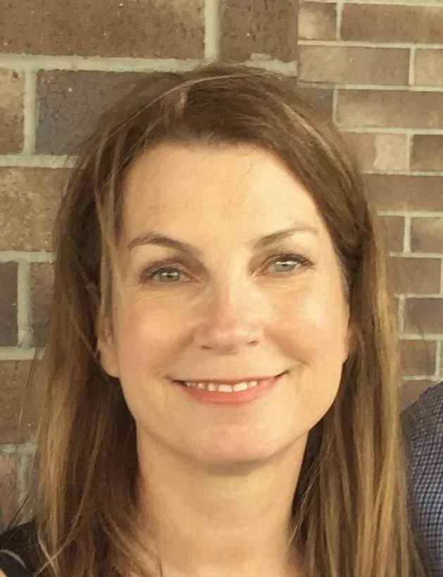 A woman with light brown hair, smiling, standing in front of a brick wall.