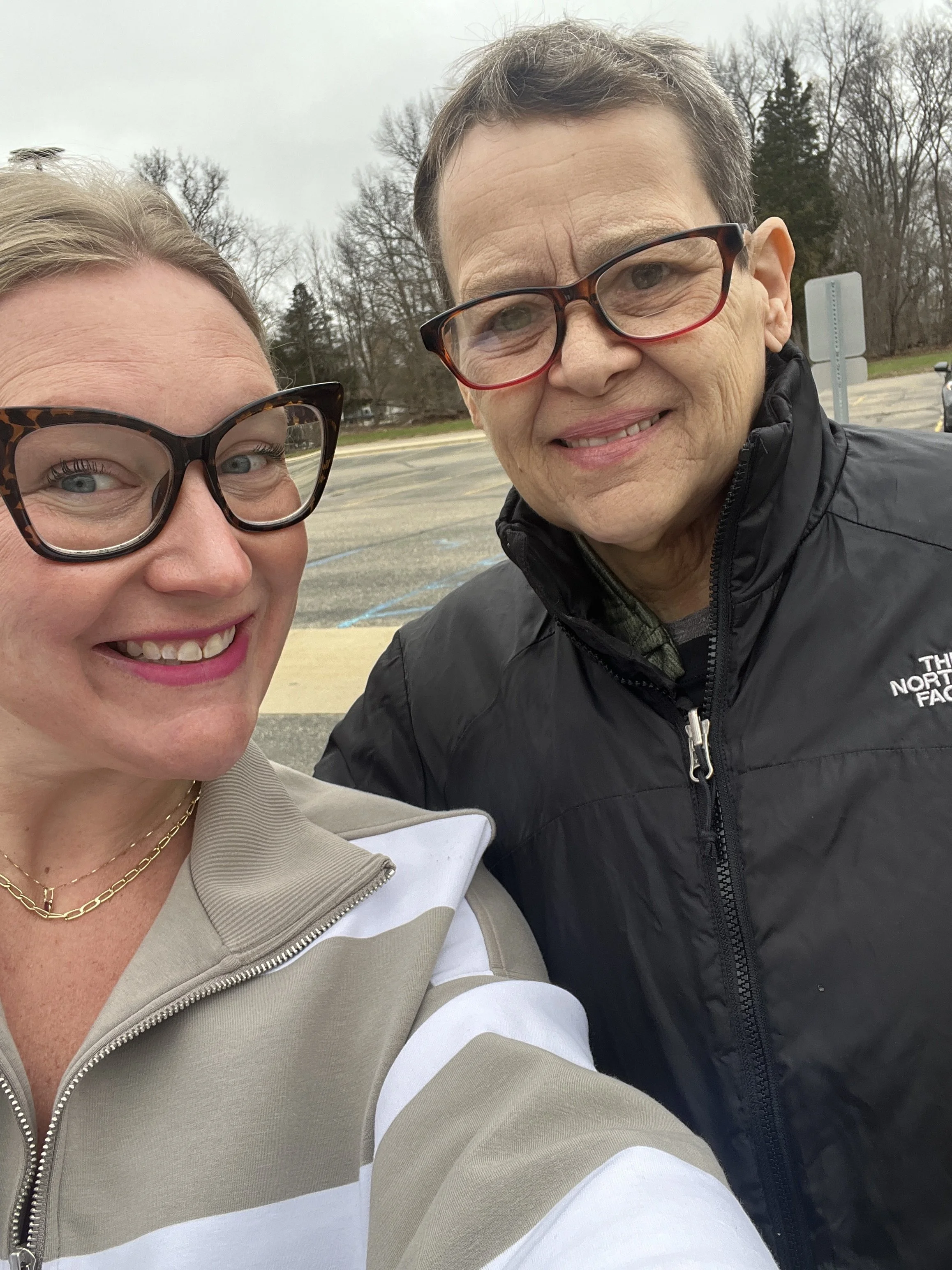 Two women taking a selfie outdoors on a cloudy day, with trees and a parking lot in the background. The woman on the left has blonde hair, is wearing large black tortoise shell glasses, a light-colored jacket with a zipped front, and gold necklaces. The woman on the right has short dark hair, is wearing red-rimmed glasses, and a black North Face jacket.