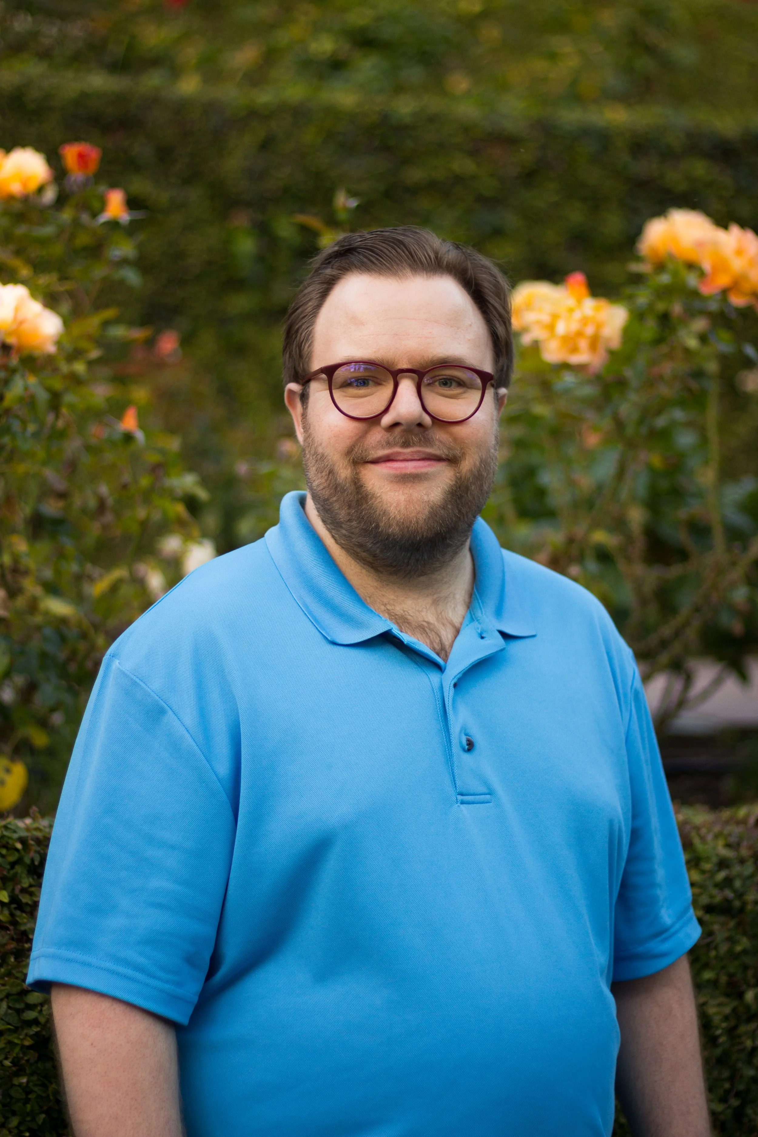 a bearded white man with light brown hair and glasses wearing a blue collared shirt stands in front of roses