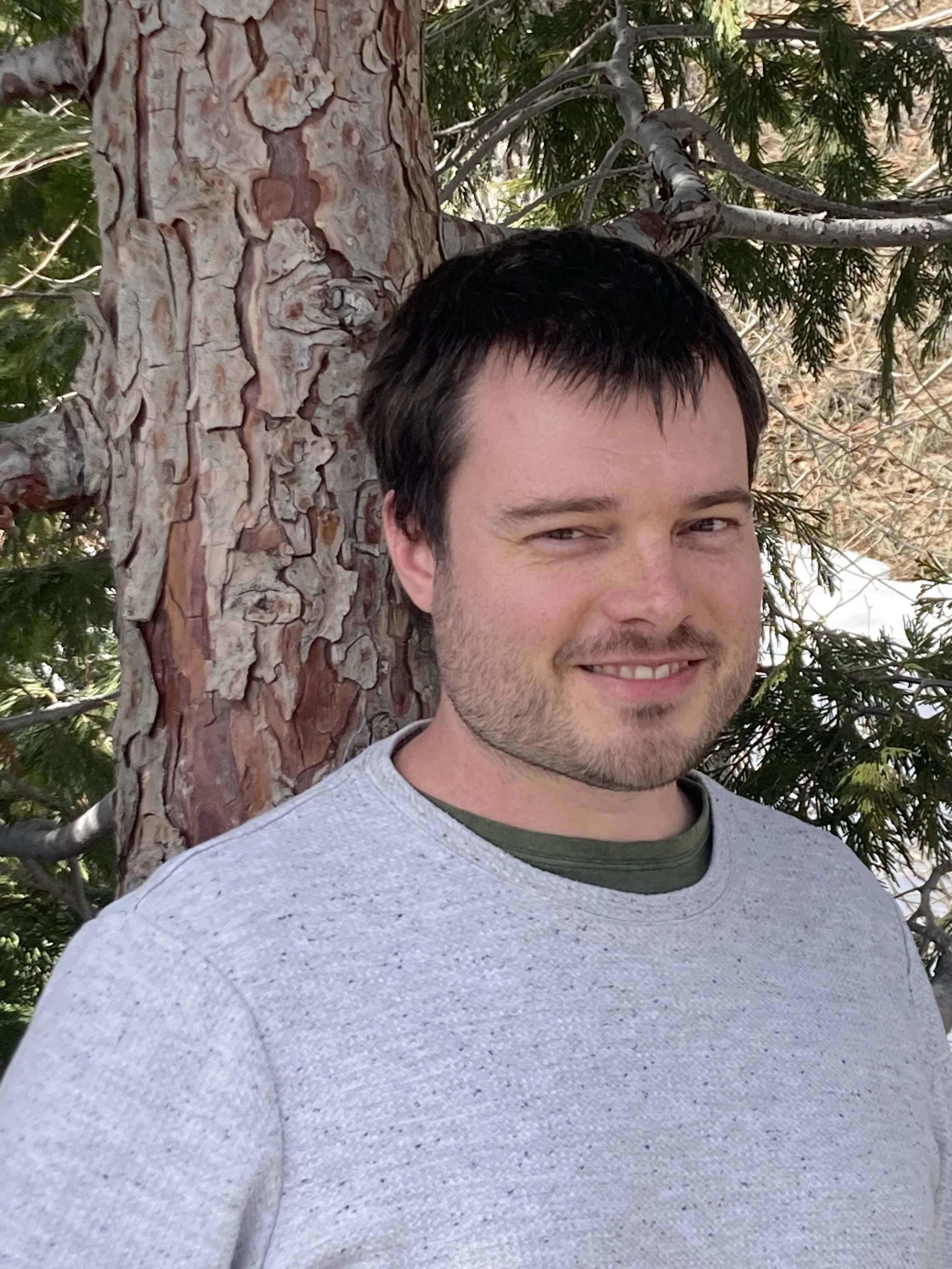 a young white man with scruffy facial hair smiles in front of a tree.