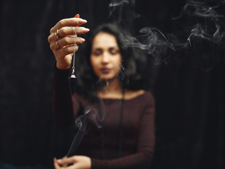 Woman holding a pendulum necklace with smoke rising around her against a dark background.