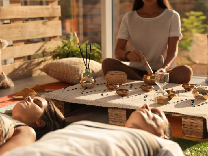 A woman receiving a crystal healing massage while lying on a bed outside, with another person lying nearby. A person is meditating or doing a sound therapy session at a low table with bowls, an hourglass, and candles.