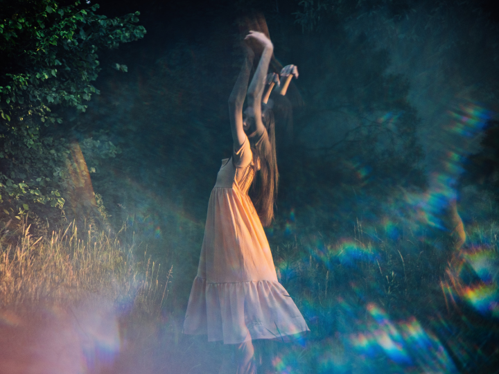 A girl in an orange dress standing outdoors surrounded by trees and grass, with light effects creating rainbow-like lens flares.