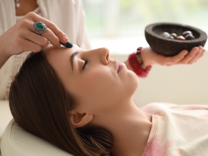 A woman receiving a facial treatment, with a practitioner applying a facial mask or cream with a brush on her forehead. The woman has her eyes closed and appears relaxed.
