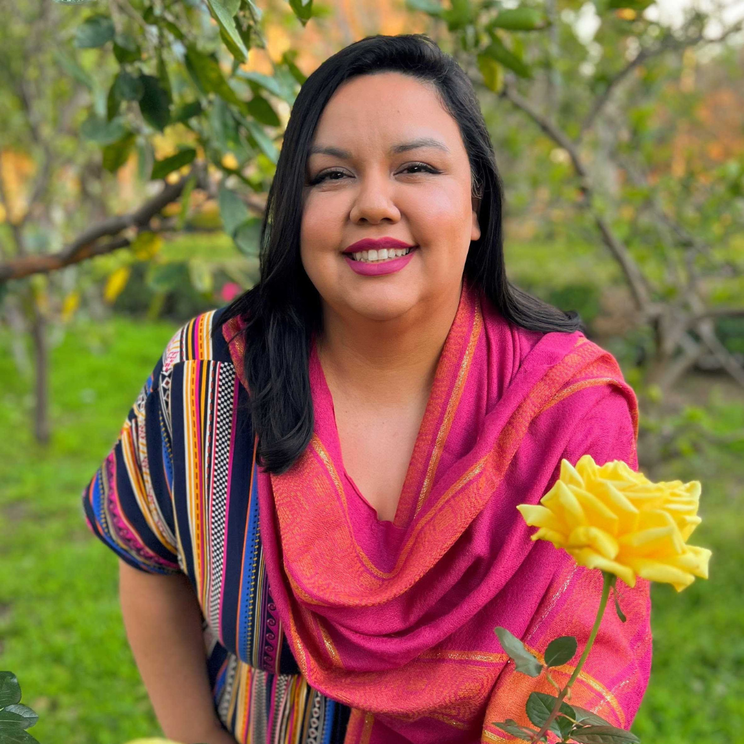 A woman with black hair and pink lipstick smiling outdoors, wearing a colorful, striped shirt and a pink scarf, with yellow roses in front of her, green trees and grass in the background.