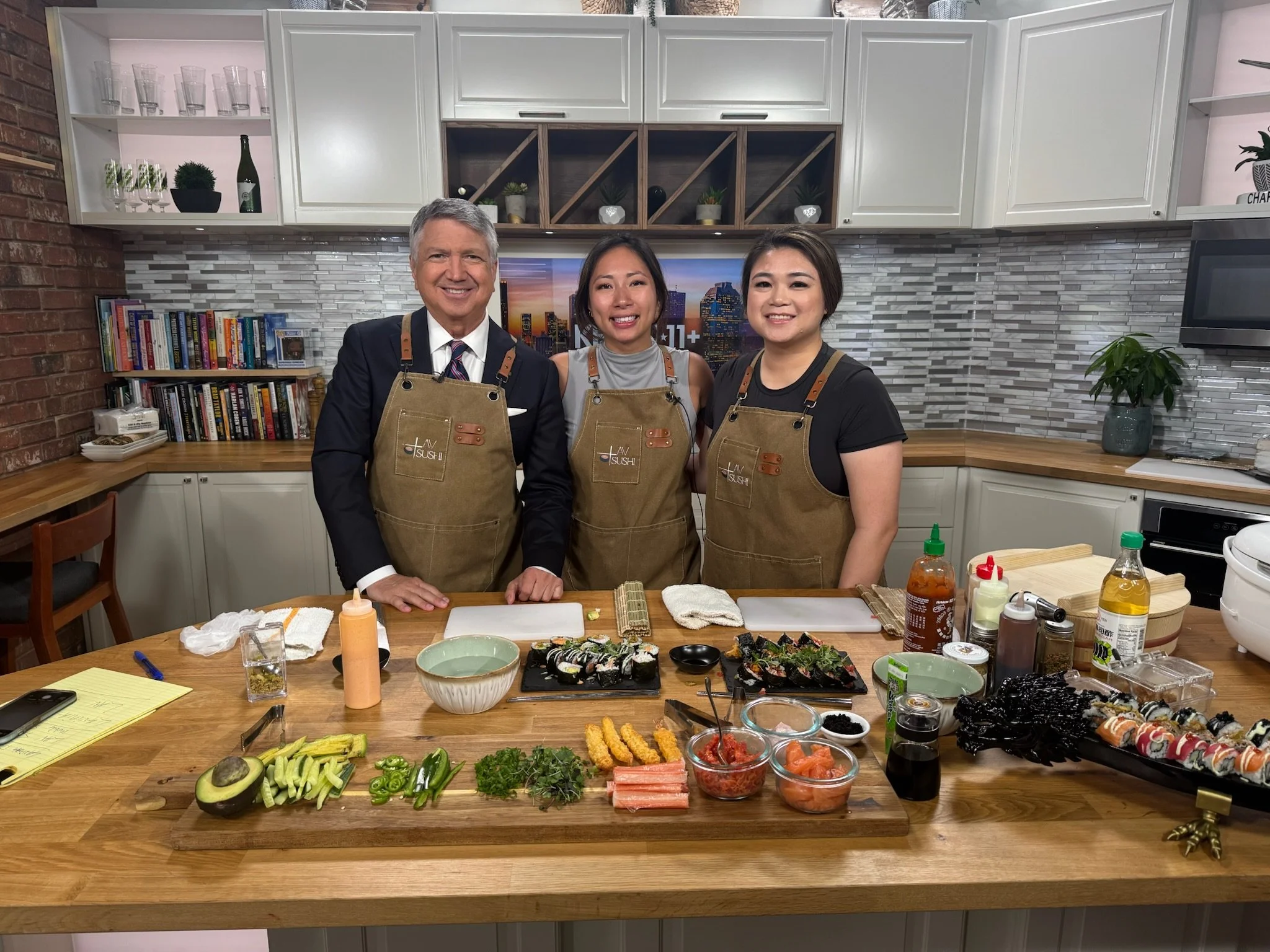 Three people wearing aprons standing behind a sushi-making counter in a modern kitchen with a variety of sushi ingredients and rolls on the table.