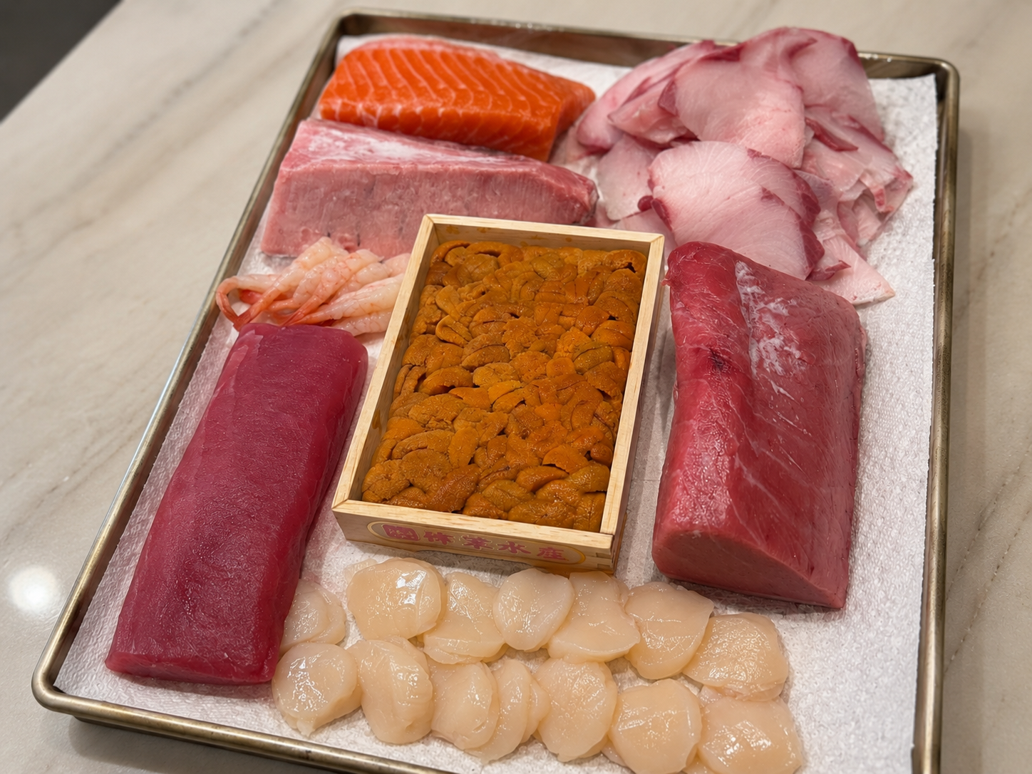 Assorted fresh seafood and fish roe on a metal tray, including slices of tuna, salmon, and other fish, shrimp, sea urchin, scallops, and sashimi-grade meat.