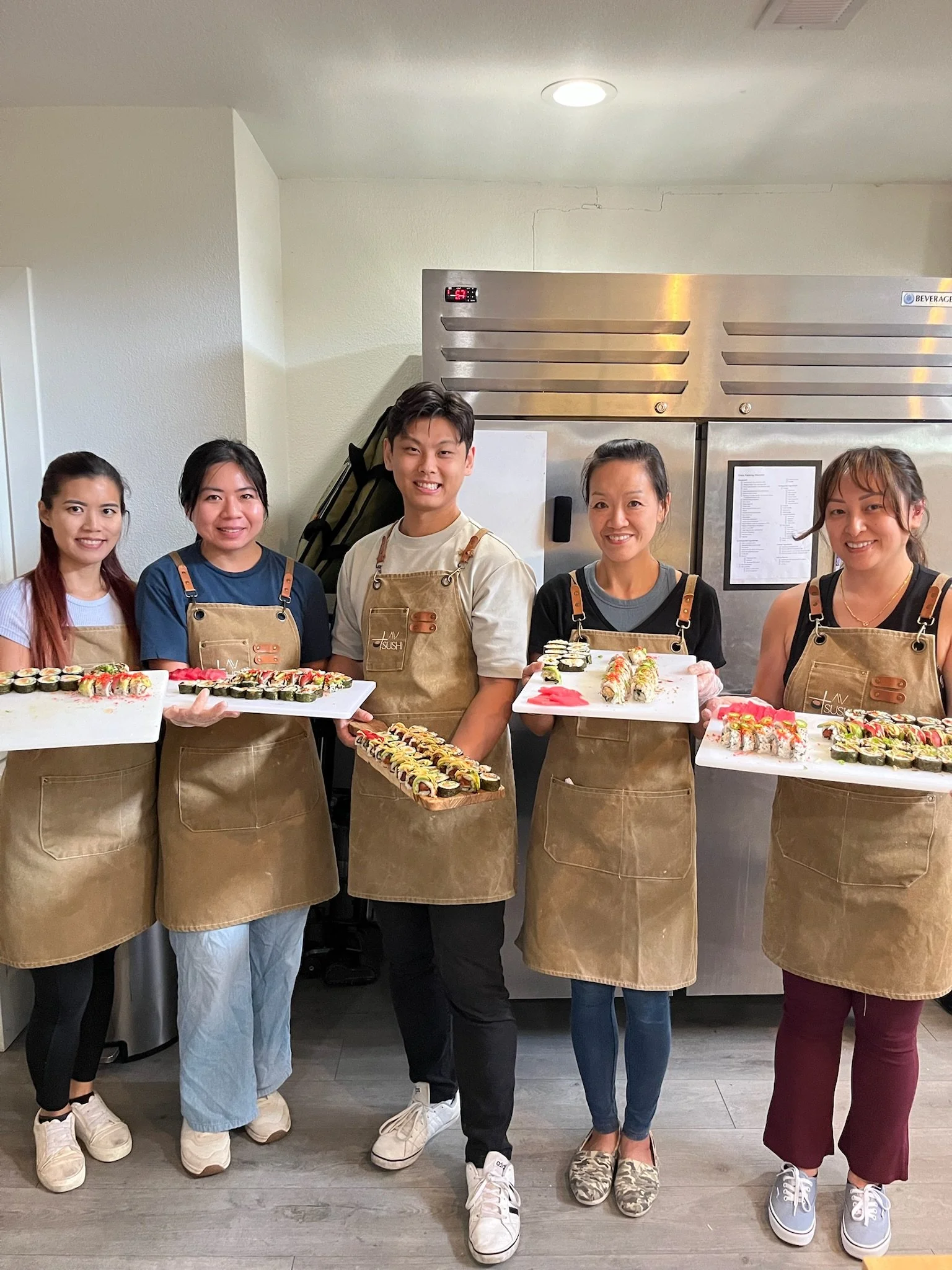 Five people standing in a row, each holding a tray of sushi, inside a commercial kitchen with white walls and stainless steel refrigerator in the background.