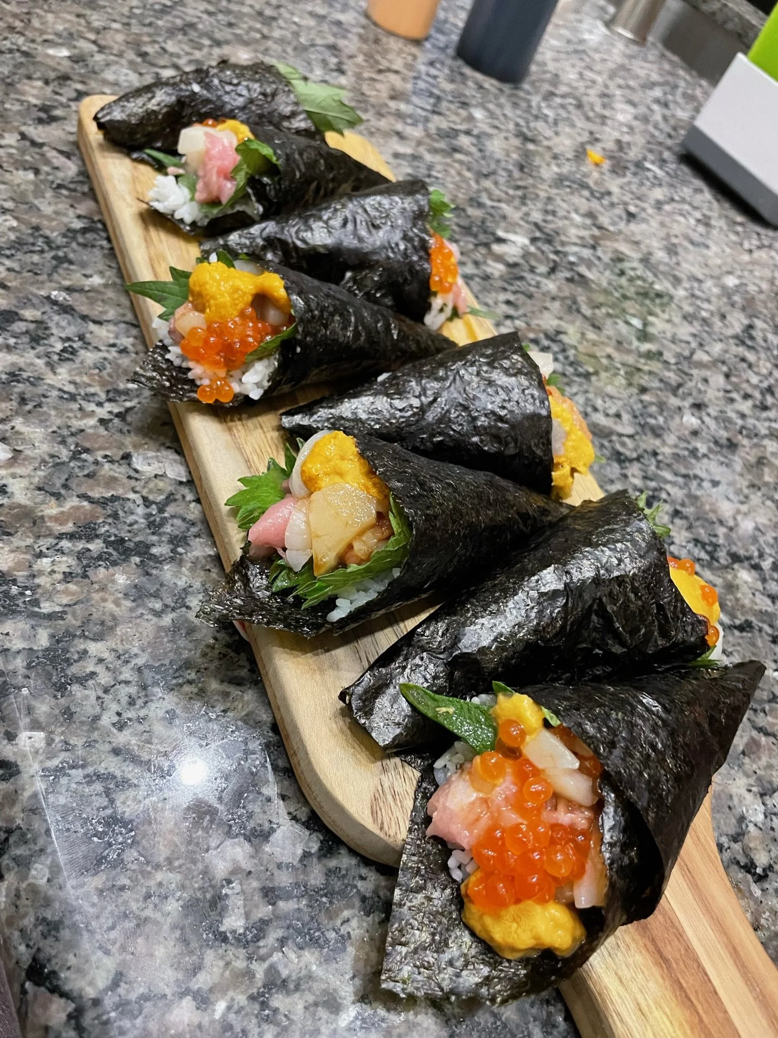 A row of sushi hand rolls with nori seaweed wraps, filled with rice, fish, and topped with various fish roe and garnishes, presented on a wooden serving board on a granite countertop.
