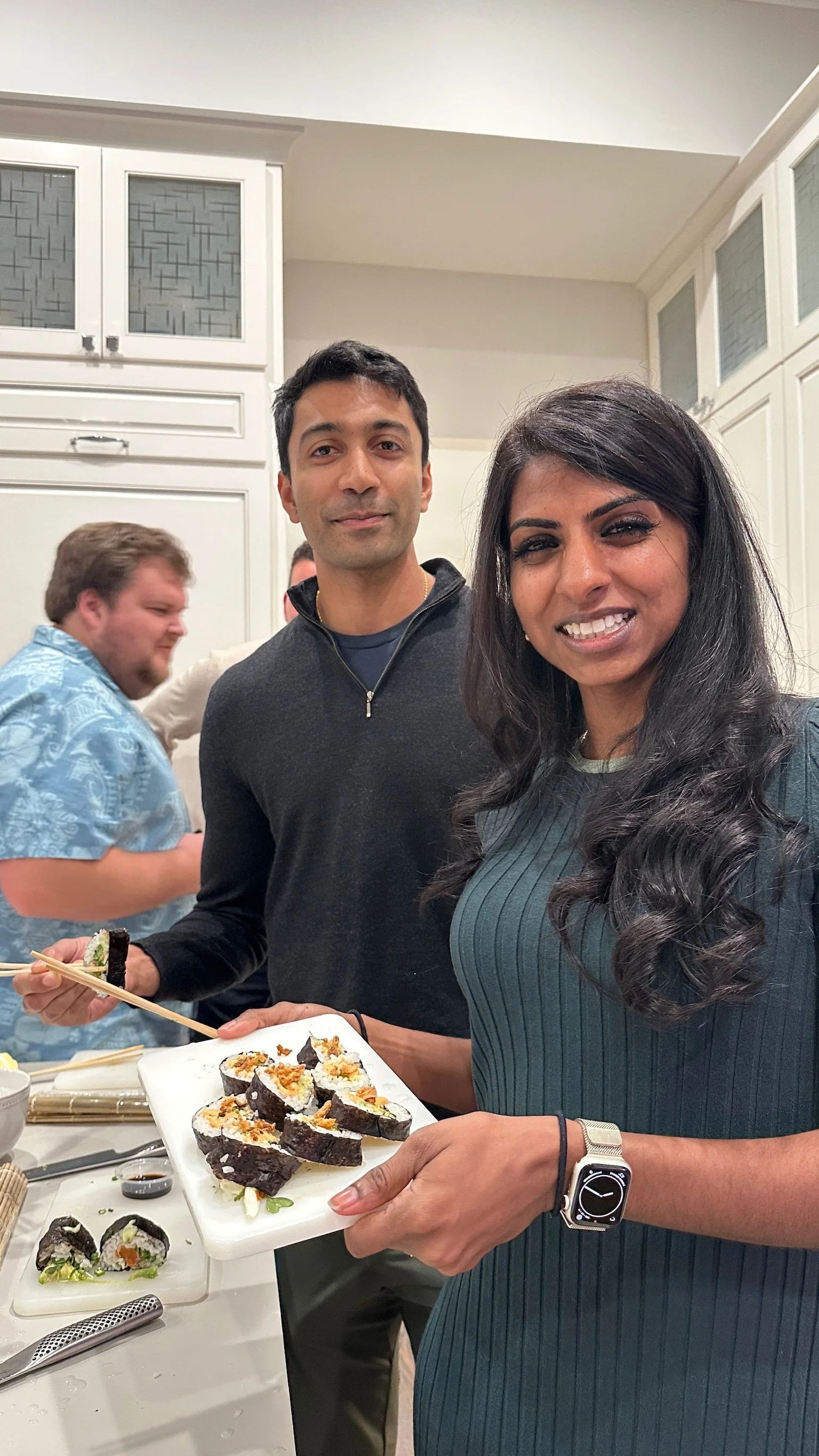 Woman with dark hair in a striped shirt holding a plate of sushi at a gathering, with two men in the background, one holding a plate and chopsticks, in a kitchen setting.