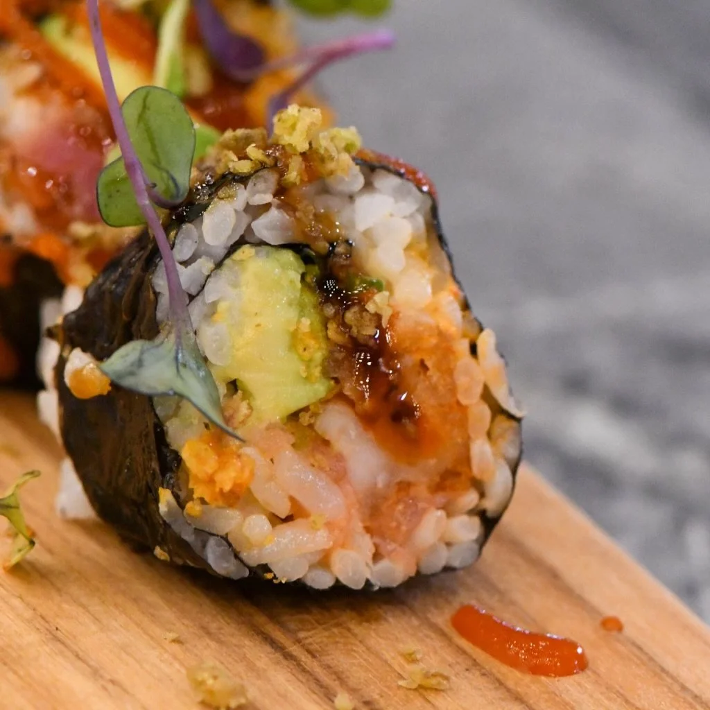 Close-up of a sushi roll with rice, avocado, cooked seafood, eel sauce, and garnished with microgreens on a wooden surface.