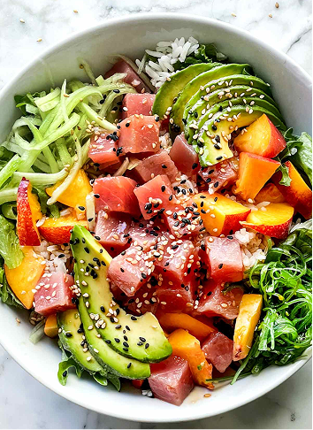 Bowl of poke salad with diced tuna, avocado slices, chopped mango, rice, and mixed greens, garnished with sesame seeds.