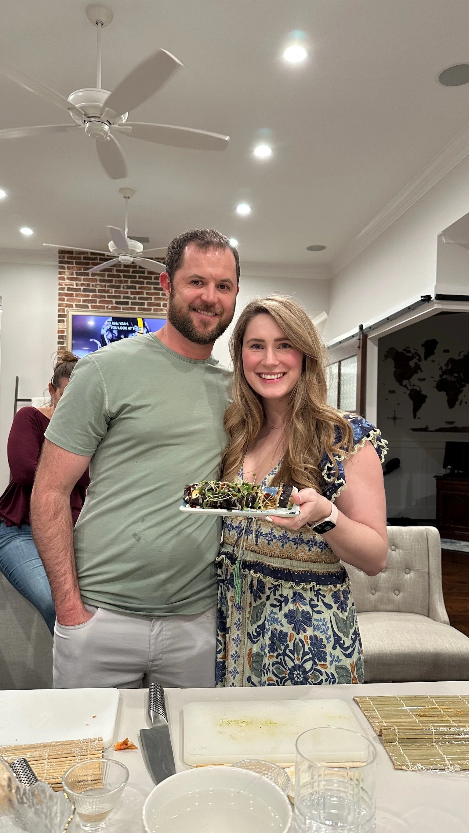 A man and a woman smiling at the camera, with the woman holding a plate of sushi. They are in a home with a brick wall and a world map on the wall in the background. There is a white ceiling fan and ceiling lights above them, and a woman in a maroon shirt is blurred in the background.