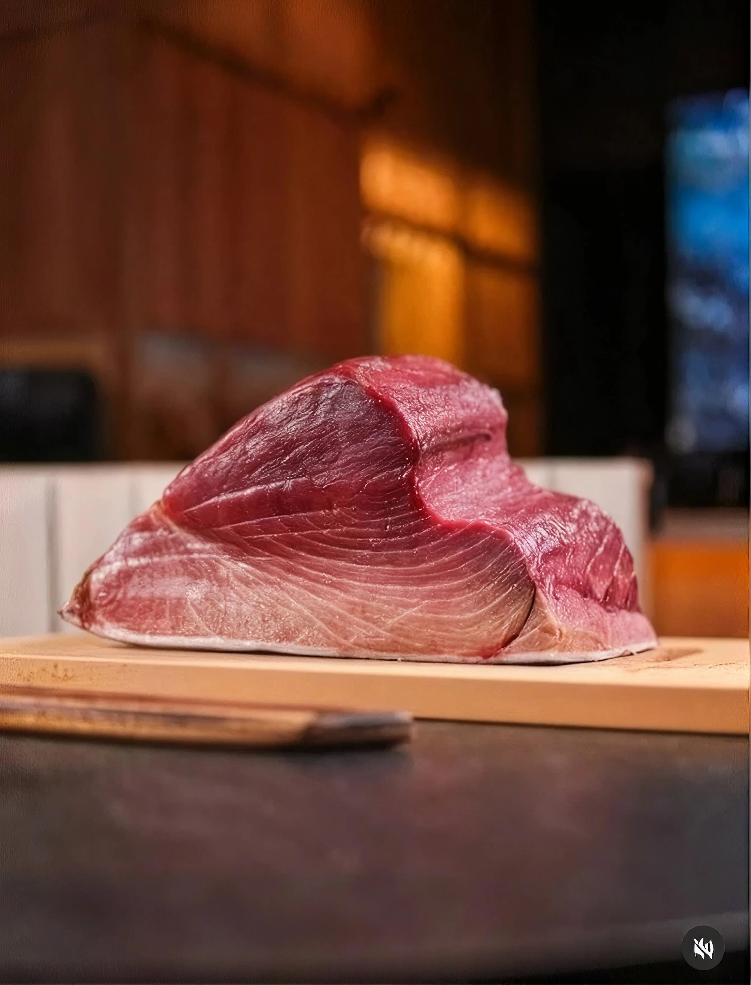 A close-up of a raw piece of red meat resting on a wooden cutting board, with a blurred kitchen background and dark surroundings.