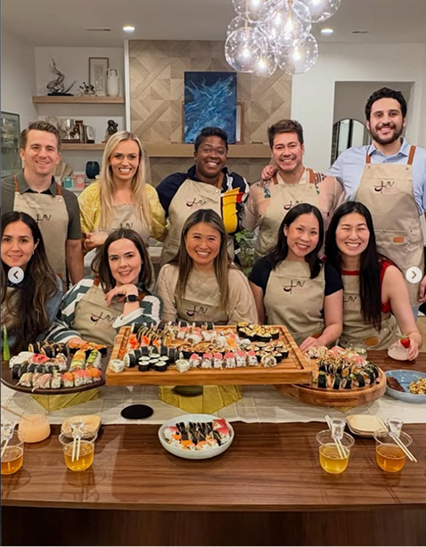 A group of nine people taking a sushi class from Lavsushi gathered around a table with sushi and drinks, celebrating together in a decorated room.