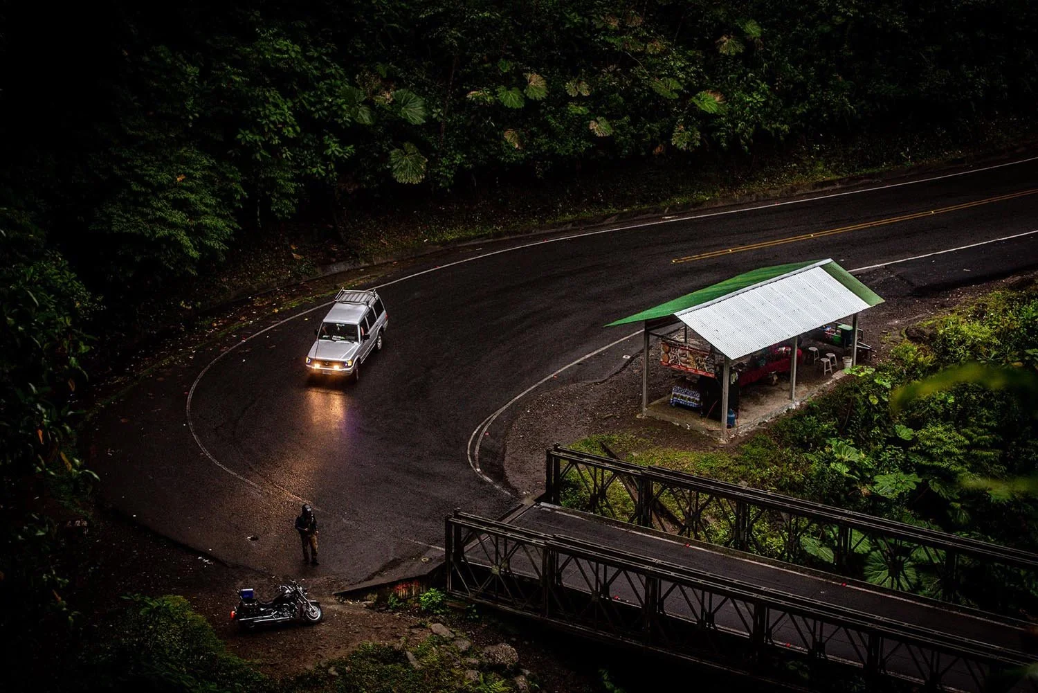 Curved road with a car and a motorcycle near a roadside stall in a lush green area.