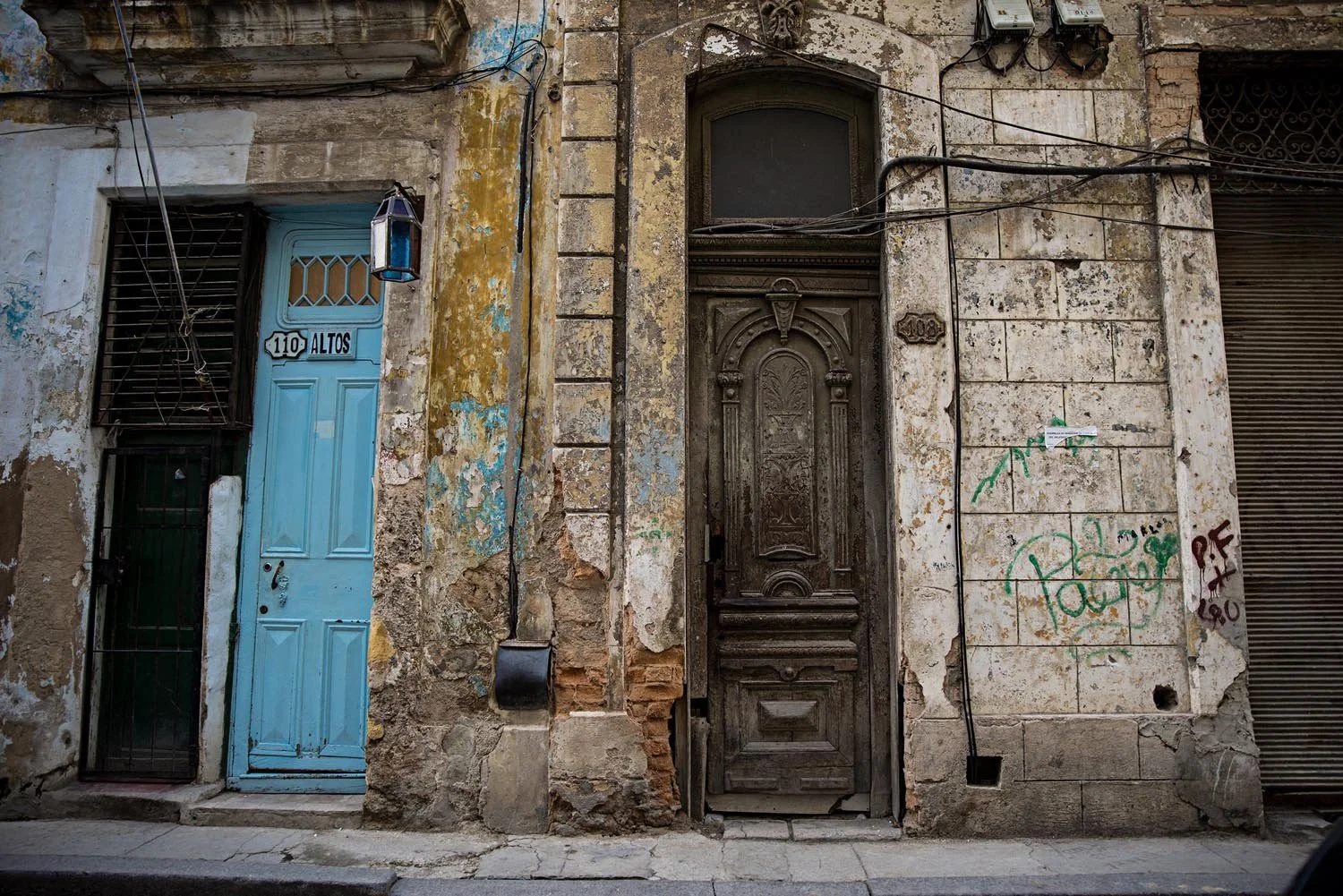 Worn urban building facade with blue and brown doors, peeling paint, graffiti, and an old lamp.