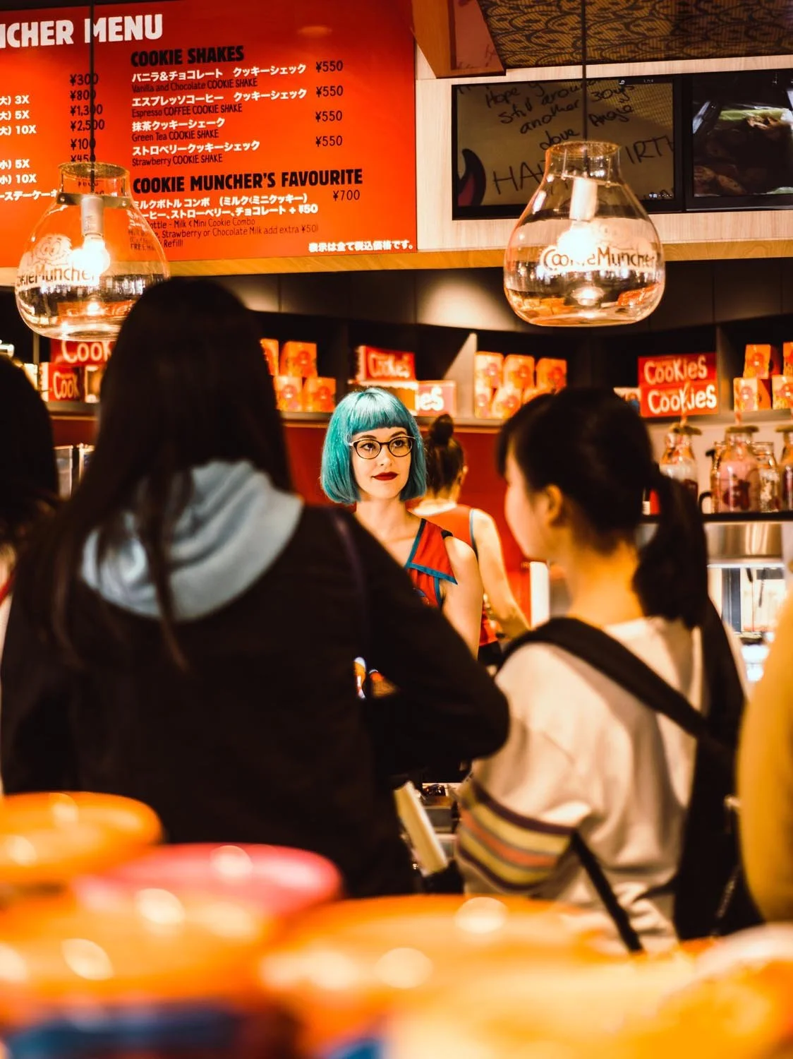 A woman with blue hair and glasses stands behind a counter at a cookie shop, assisting customers. The shop has a menu board with prices for cookie shakes, along with colorful packaging and lamps.