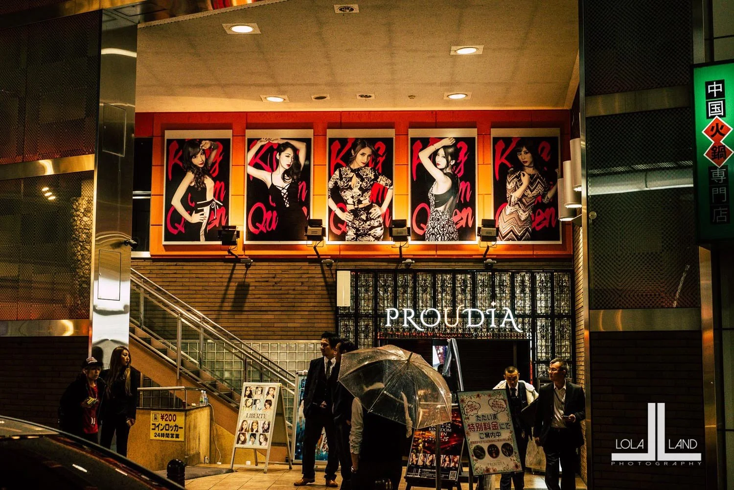 Nighttime street view of a building entrance in Japan, featuring illuminated posters of women on the facade, people standing outside with umbrellas, and signage for "PROUDIA" above the entrance.
