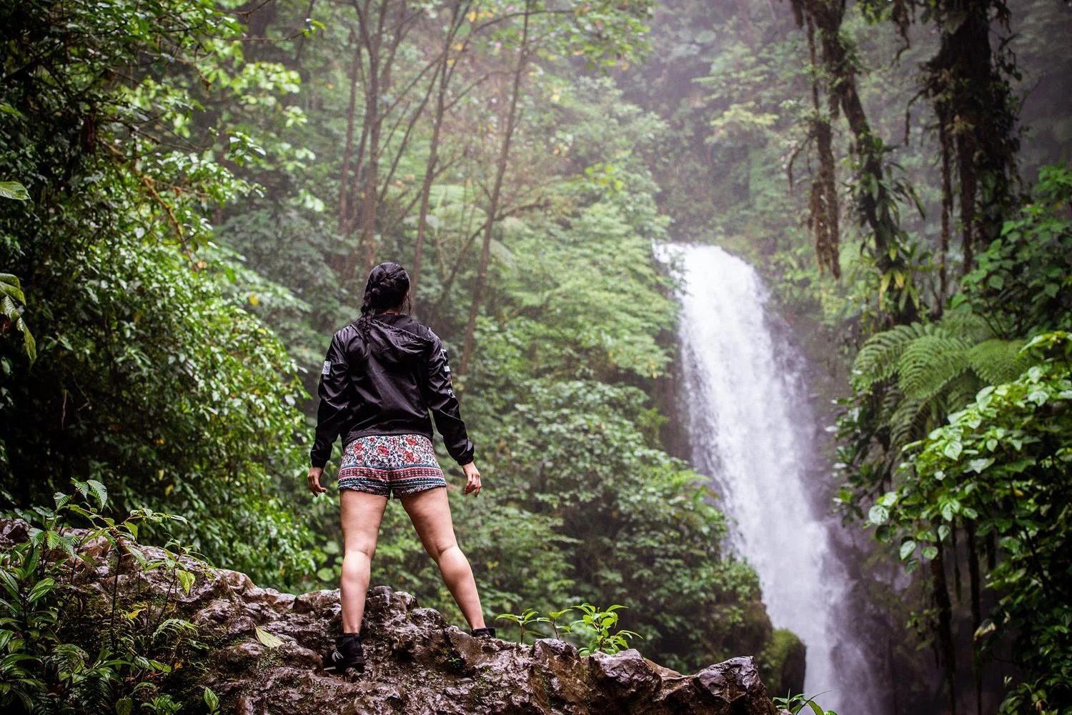 Person standing on rocky terrain gazing at a waterfall surrounded by dense green foliage in a forest setting.