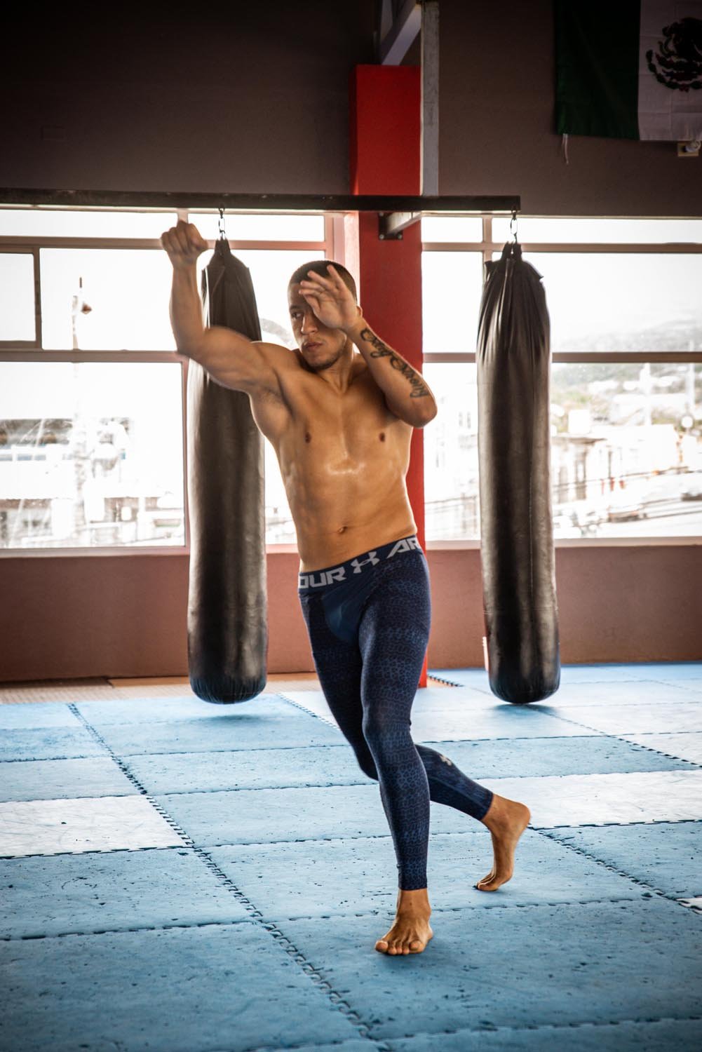 Man practicing martial arts in a gym with punching bags and mats, wearing workout leggings.
