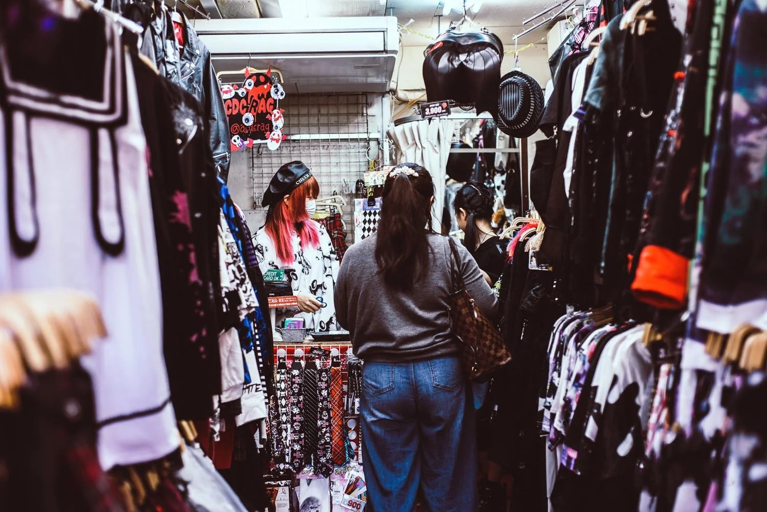 Shoppers in a clothing store with various clothes hanging, a cashier with pink hair and a mask standing behind the counter.