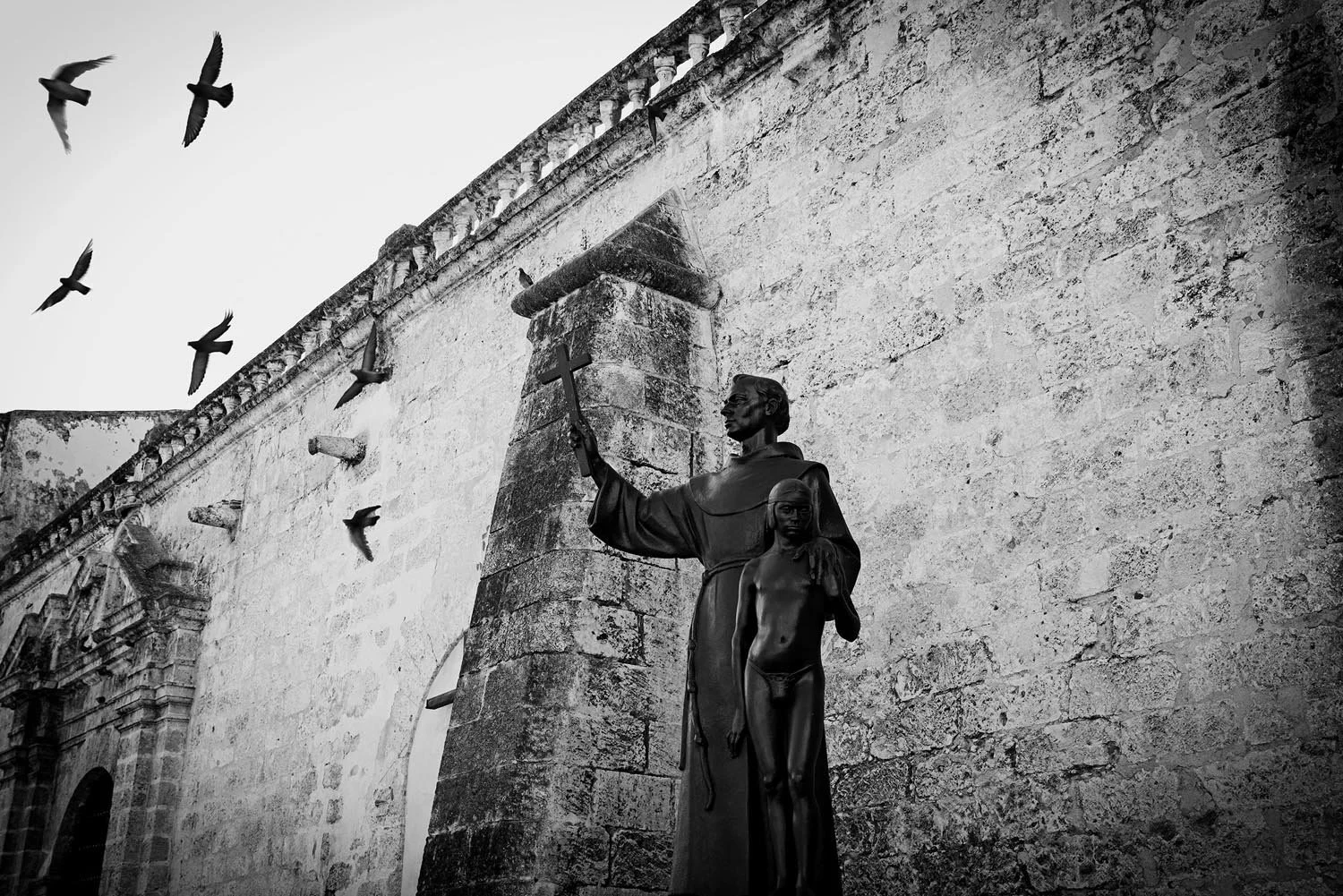 Black and white photo of a historical stone building with a statue of a religious figure holding a cross. Birds are flying nearby.