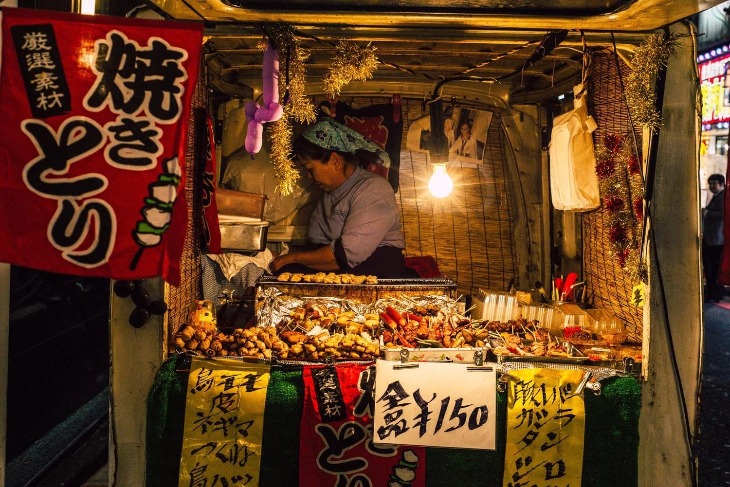 Japanese street food stall with yakitori skewers and handwritten price signs, vendor inside, illuminated by a hanging light bulb, decorated with colorful banners.