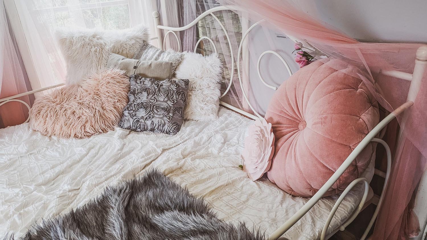 Cozy bed with metal frame adorned with fluffy and decorative pillows in pink, gray, and beige tones, and a sheer pink canopy curtain.