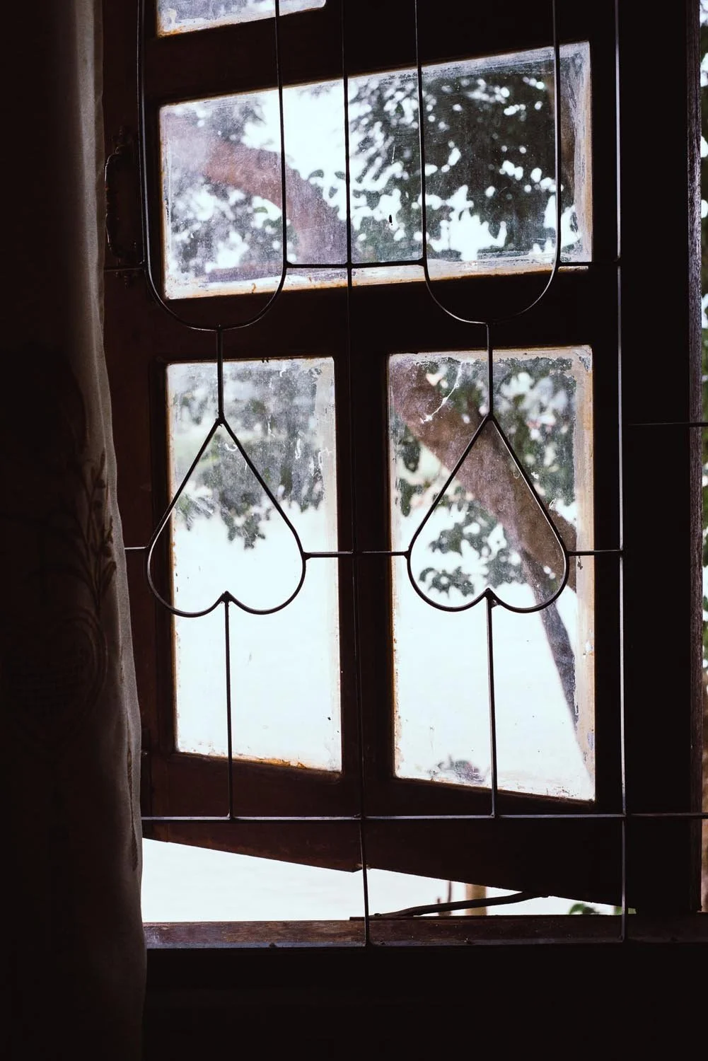 Close-up of a wooden window with a metal grid featuring heart shapes, showing a blurred view of trees outside.