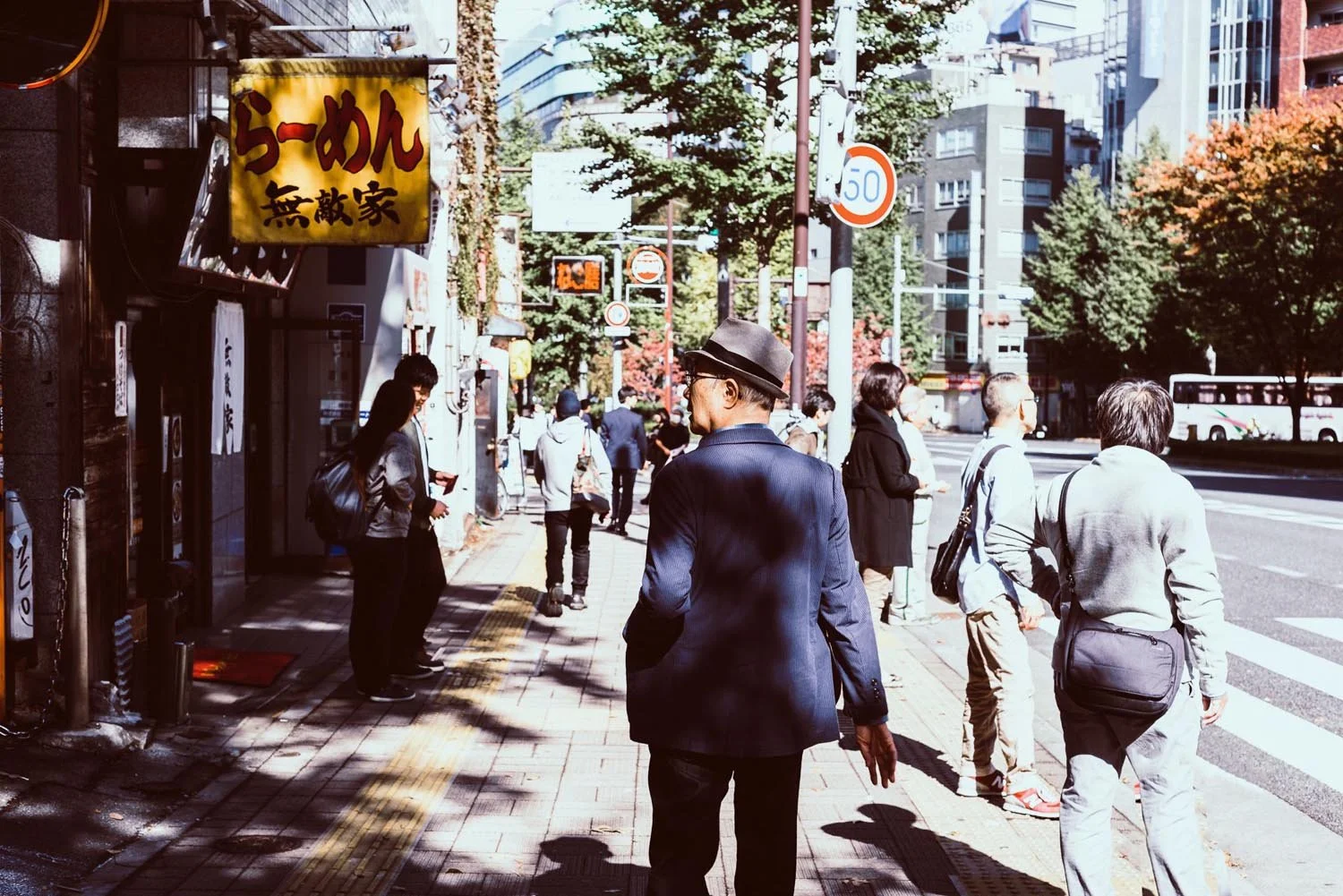 People walking on a city sidewalk in Japan near a yellow sign with Japanese characters, trees and buildings in the background.