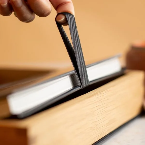 Hand pulling black ribbon bookmark from book with wooden edges.