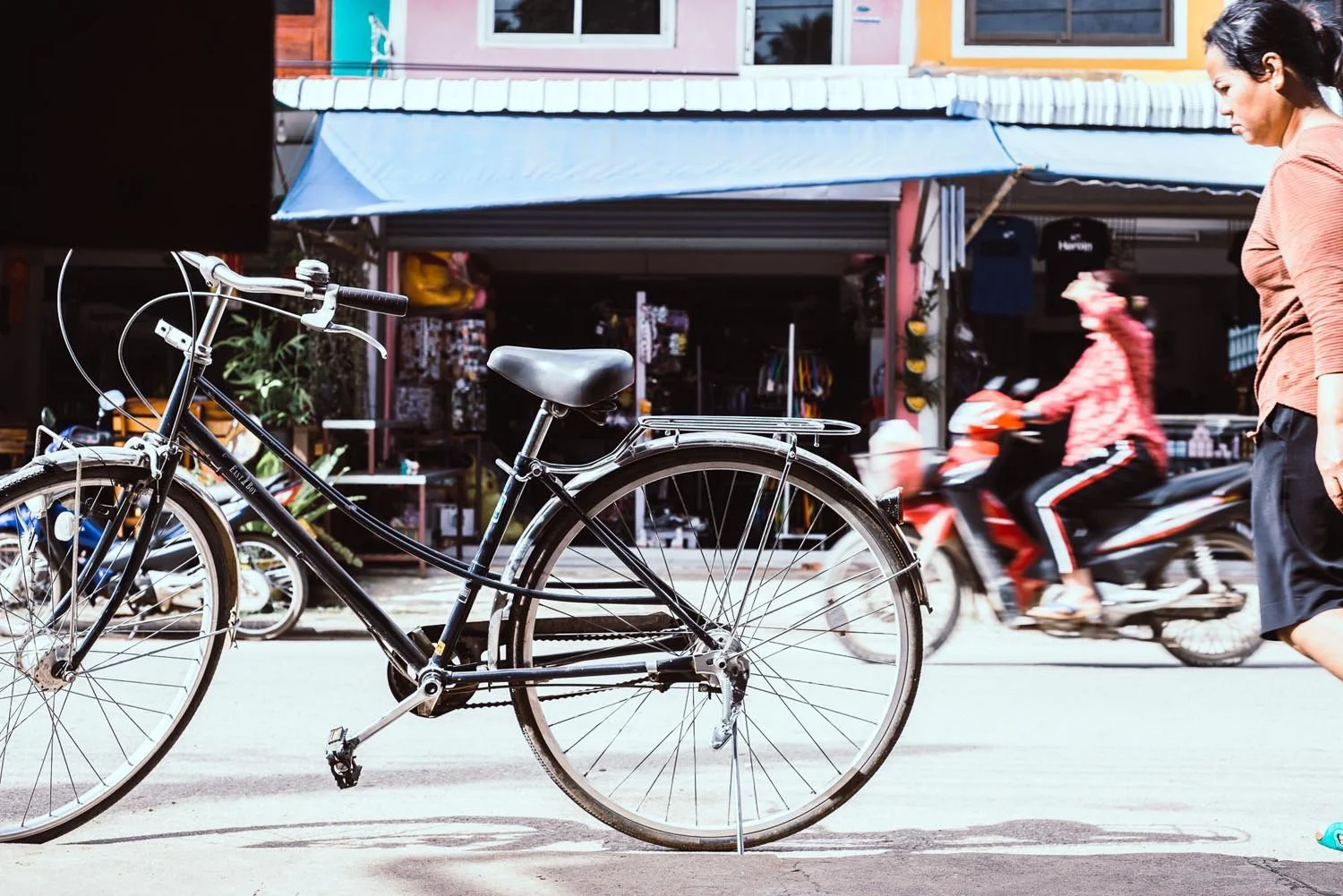 A black bicycle parked on a street with a woman walking by and a motorbike in motion in the background. Shops are visible on the other side of the street.