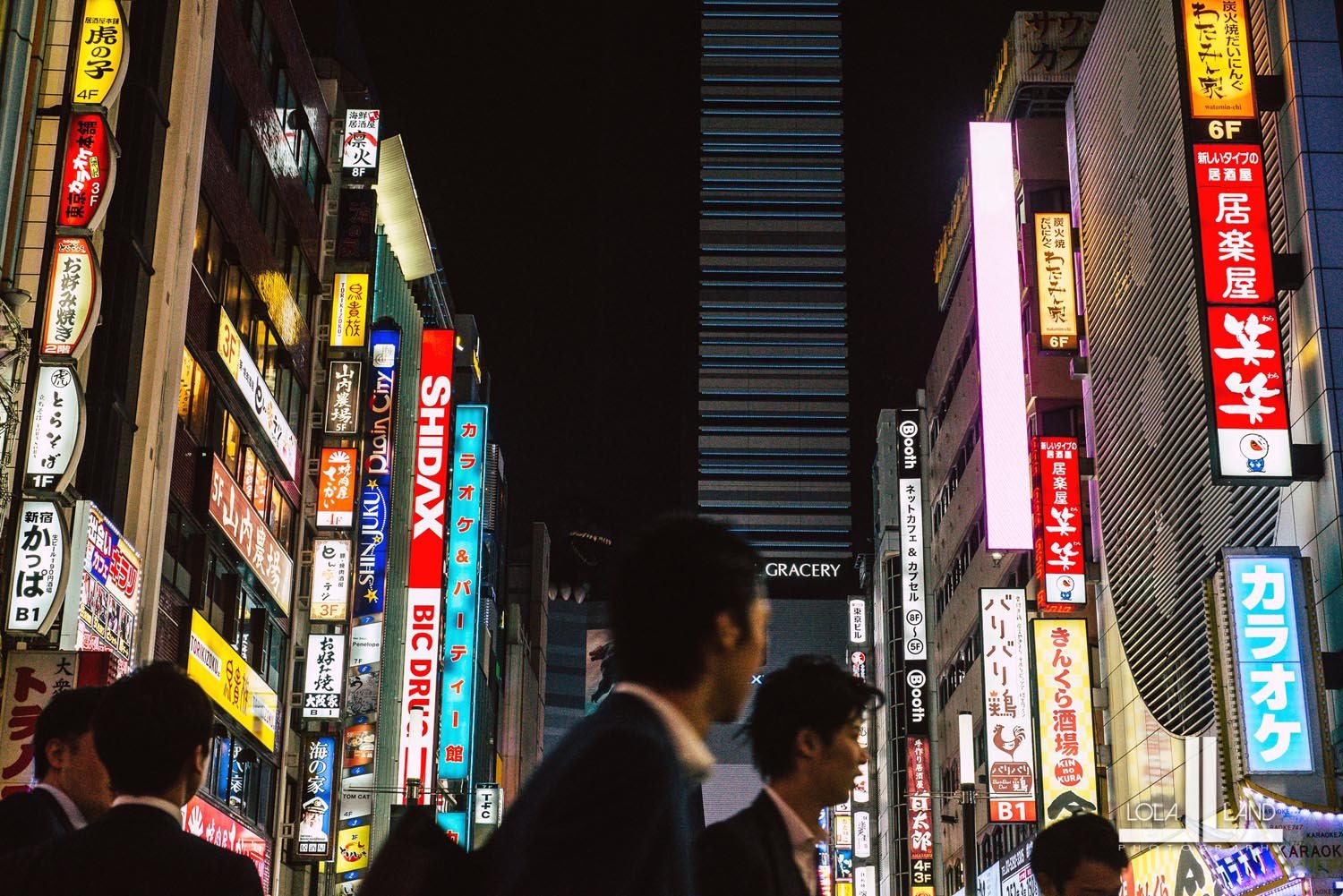 Night scene in a busy Japanese city with illuminated signs on buildings and silhouettes of people walking.