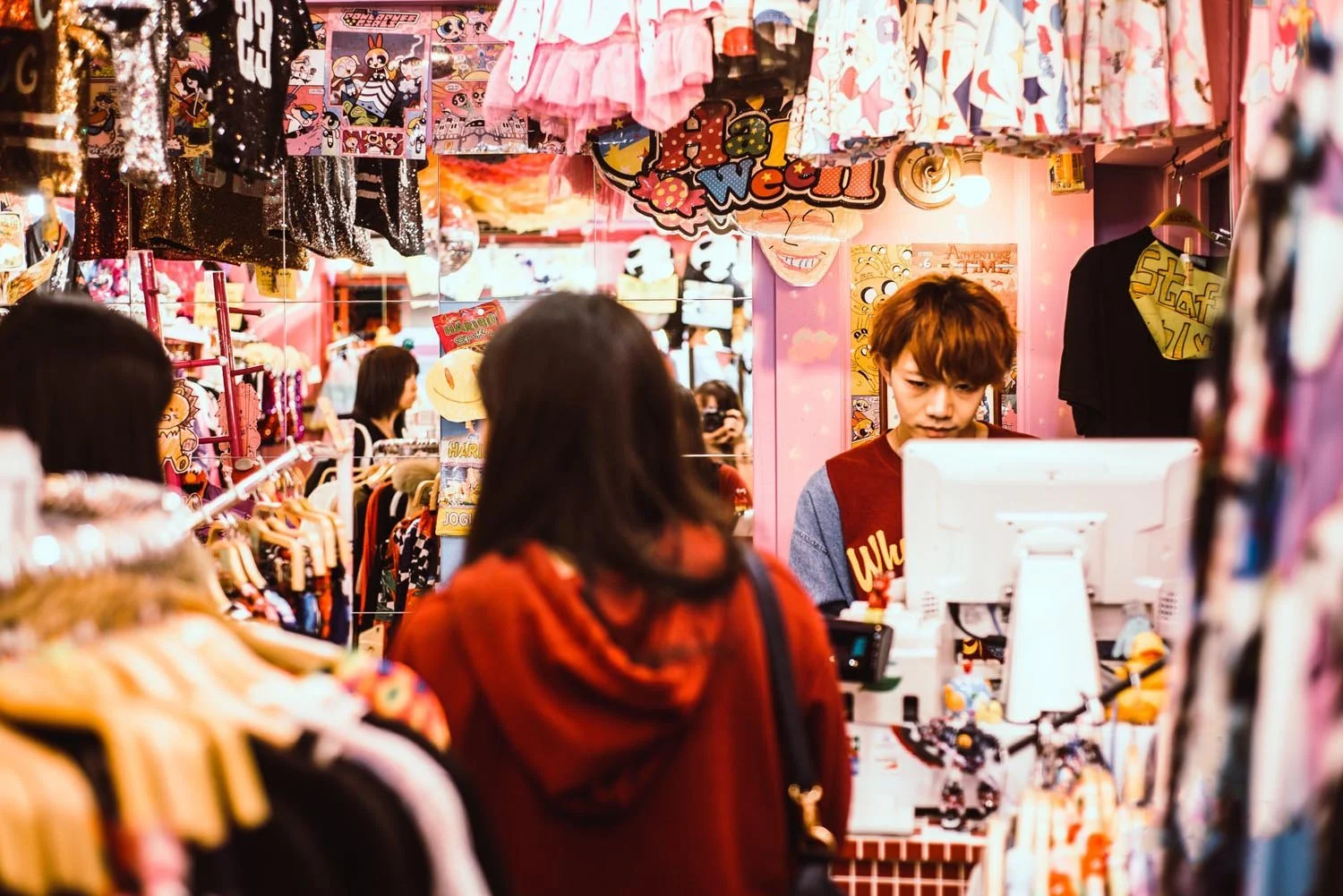 A busy clothing store with a cashier behind a register, surrounded by racks of colorful clothing and cartoon-themed decorations.