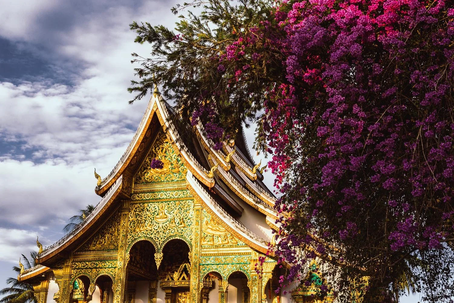 Ornate temple with intricate golden designs, surrounded by purple bougainvillaeas, against a cloudy sky.