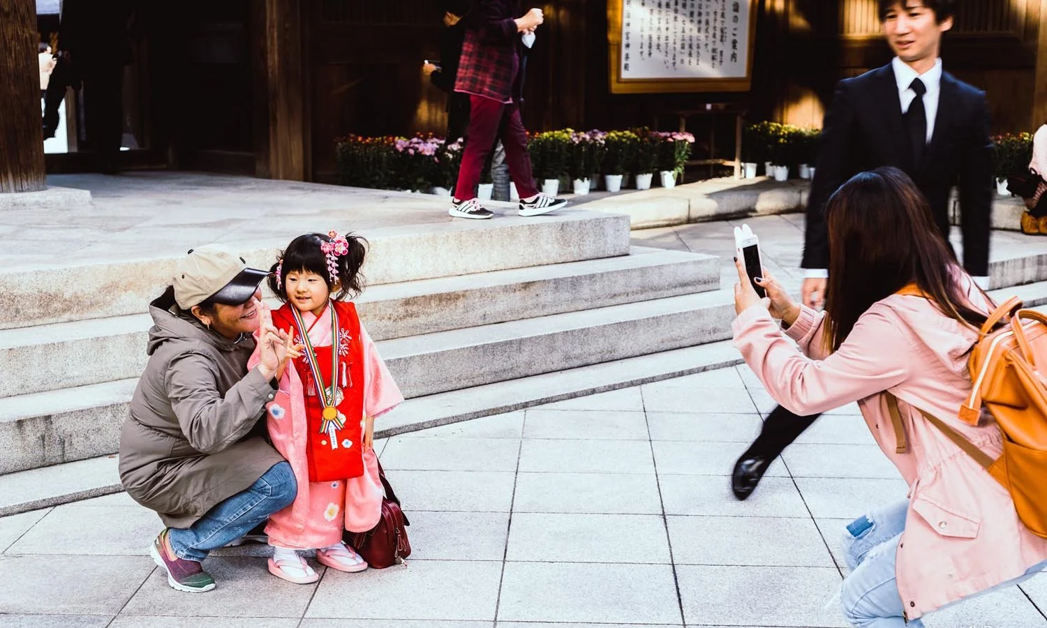 A young girl in a traditional kimono being photographed in a public setting. A person is kneeling beside her, and another is taking a picture with a smartphone. A man in a suit is walking past.