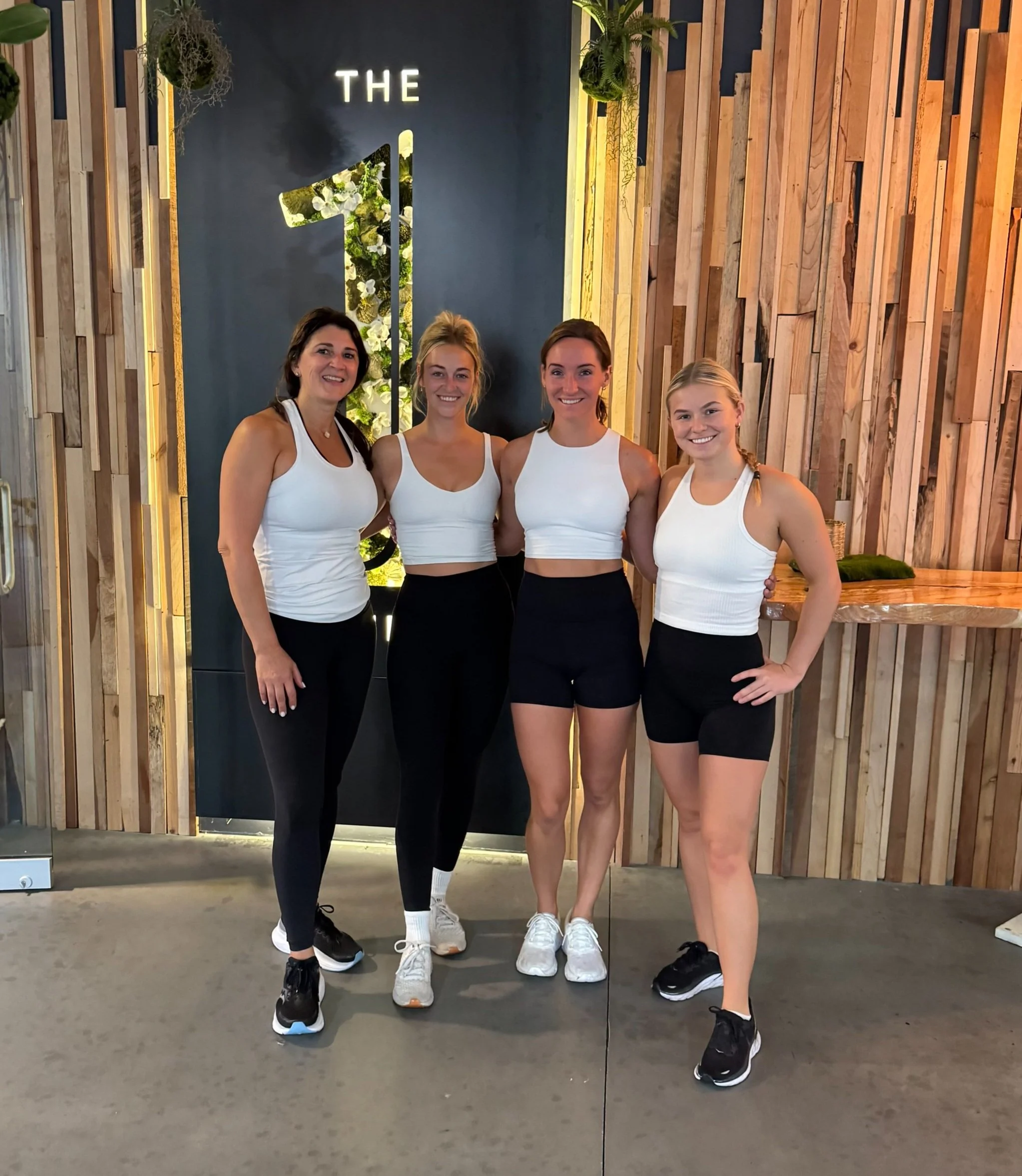 Four women in exercise attire standing together in front of a wall with large illuminated sign reading 'THE 1' and a decorative floral design, with wooden wall paneling on the right side.