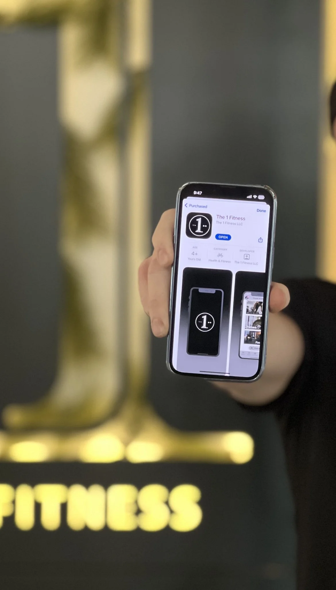 Person holding a smartphone displaying "The 1 Fitness" app with a blurred background of a fitness logo.