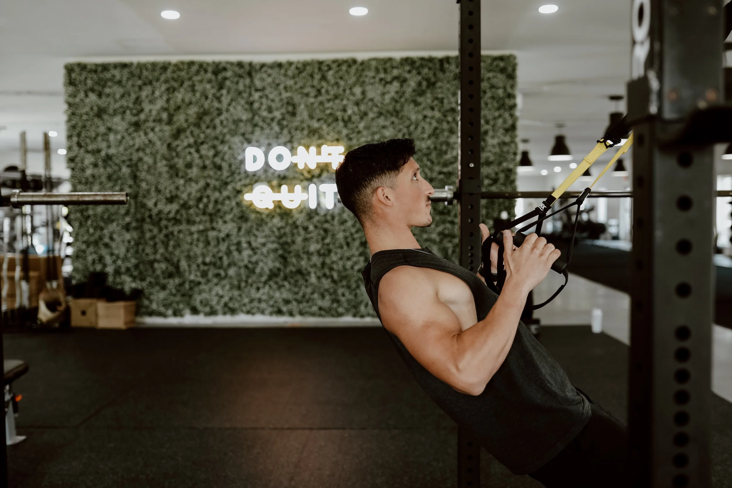 A man using suspension training straps in a gym with a green leafy wall and neon sign in the background.